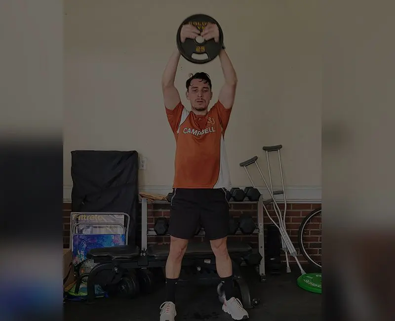 A man lifts a weighted ball above his head in a home gym.